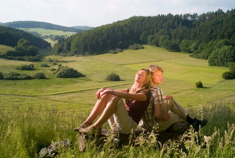 Zwei Personen sitzen auf einer Bank in einer grünen Landschaft mit Hügeln und Wäldern im Hintergrund.