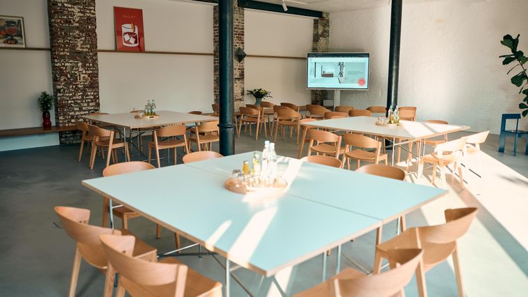 A modern conference room with several tables and chairs. A screen is mounted on the wall, and the lighting feels friendly and welcoming.
