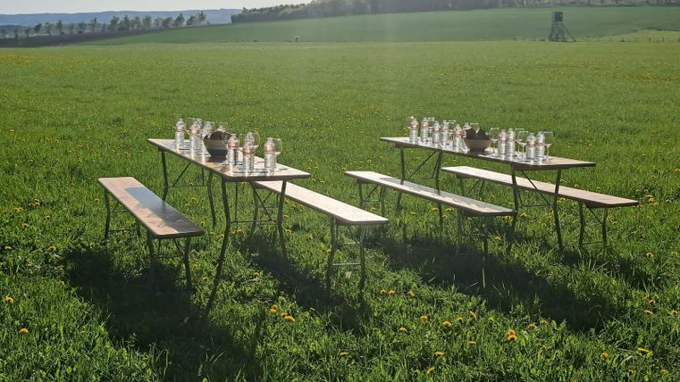 Ein gedeckter Tisch auf einer Wiese mit Getränken und Holzbänken. Im Hintergrund sind Wälder und eine weite Landschaft zu sehen.