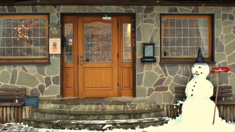 Ein holzverkleidetes Haus mit einer steinernen Fassade und großen Fenstern. Draußen steht ein Schneemann mit einem schwarzen Hut und einer roten Schaufel.