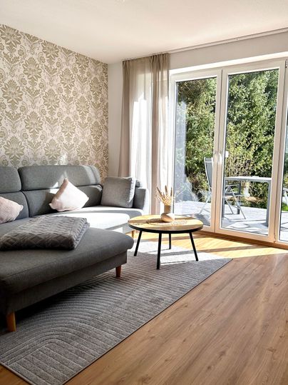 Een gezellige woonkamer met een grijze bank en een kleine tafel. Grote ramen laten veel licht binnen en bieden uitzicht op het groen.