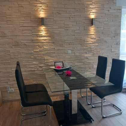 Een moderne eetruimte met een glazen tafel en zwarte stoelen. De muren zijn versierd met steenplaten en twee wandlampen zorgen voor een gezellige sfeer.