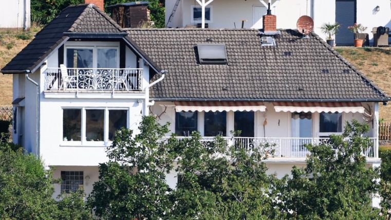 Een modern huis met een balkon en een schuin dak. Omgeven door bomen en een lichte omgeving.