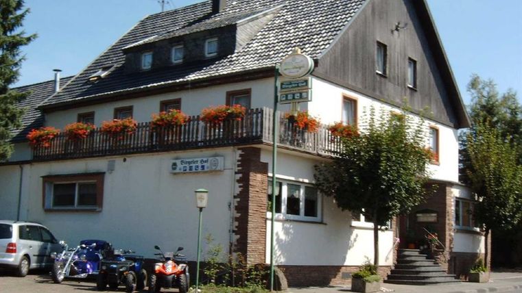 <p>Een drie verdiepingen tellend gebouw met een zwart dak. De ingang is te bereiken via een trap. Op de eerste verdieping omringt een houten leuning met bloembakken een groot balkon. Links naast het huis is een parkeerplaats, die bezet is door drie auto's. Achter de parkeerplaats hangt een bord met de tekst "Biergarten". Voor het hotel loopt een stoep die direct aan de hoofdweg grenst.</p>
