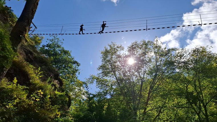 A narrow hanging bridge crosses a forest with dense trees. Two people walk on the bridge while the sun shines through the clouds.