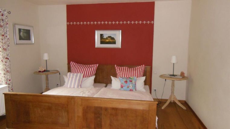 Ein helles Schlafzimmer mit einem Doppelbett und bunten Kissen. Die Wände sind in neutralen Tönen gehalten, mit einem roten Akzent und dekorativen Lampen auf Nachttischen.