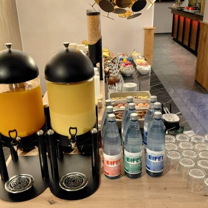 Une sélection de boissons, y compris des jus et de l'eau minérale, est disposée sur une table. En arrière-plan, on peut voir différents snacks et des verres.