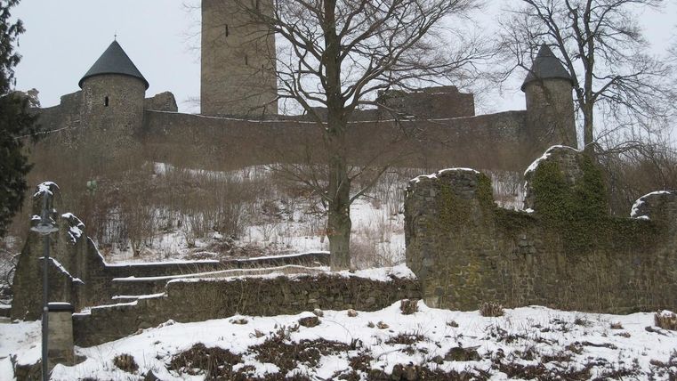An old castle ruin in the snow, surrounded by bare trees. The walls and towers are built in a historical style.