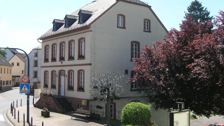 Ein Gebäude mit mehreren Fenstern und einem schönen Garten. Bäume und Sträucher umgeben die Ecke der Straße.