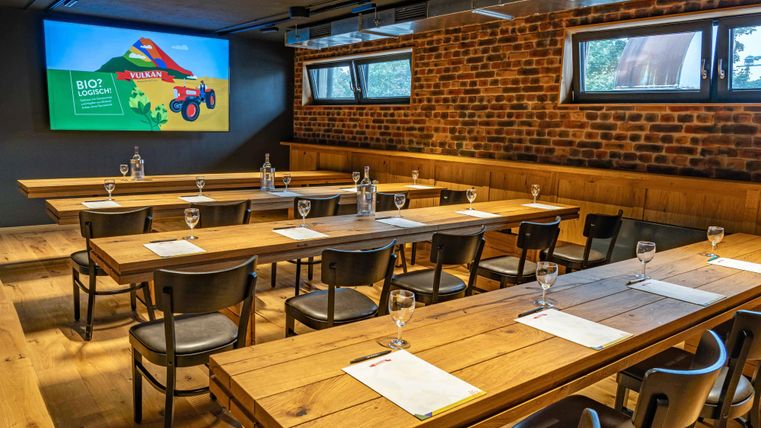 Ein moderner Konferenzraum mit langen Holztischen und Stühlen. An der Wand ist ein Bildschirm mit einer Präsentation und einer Backsteinmauer.