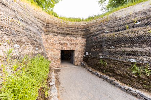 Ein Eingang aus Stein führt in einen Tunnel. Der Weg dorthin ist mit Kies bedeckt. Links und rechts stehen hohe Steinmauern. Oben wächst grünes Gras.