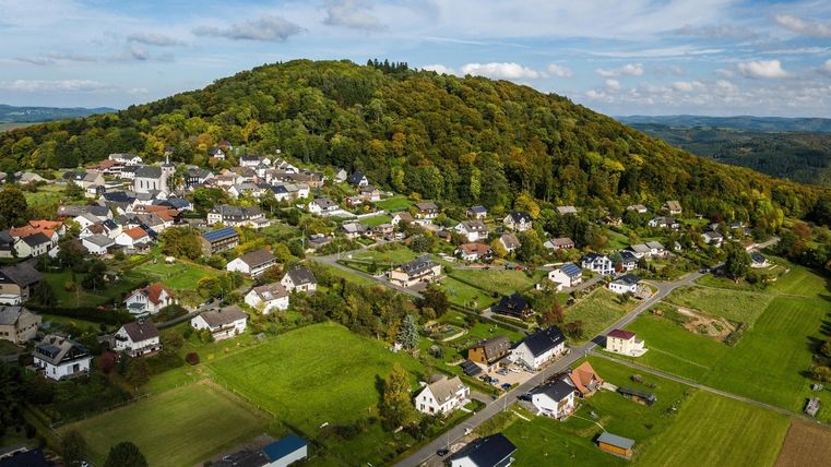 Luchtfoto van het dorp Aremberg met groene weiden en een beboste heuvel op de achtergrond.