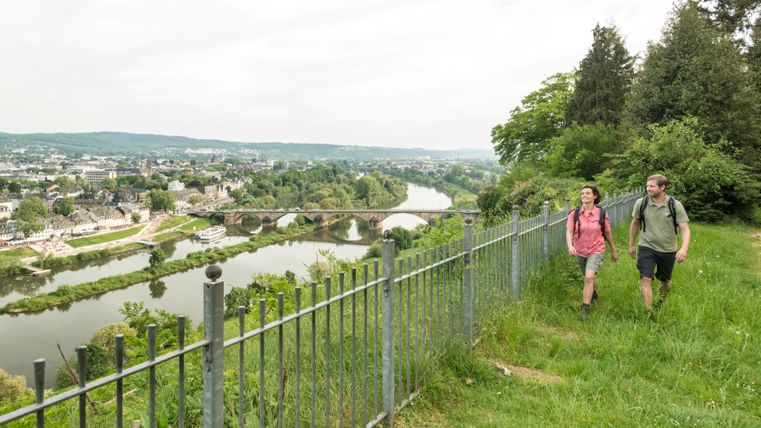 Two hikers walk along a fence with a view of the city of Trier and the Moselle.