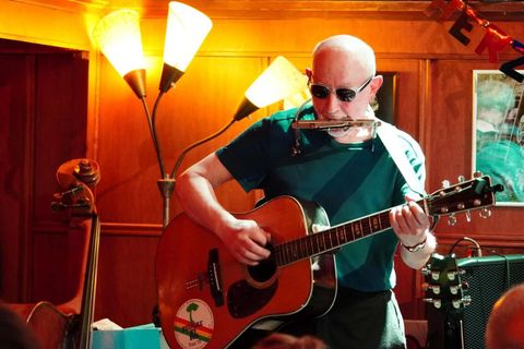 A musician plays guitar and harmonica. In the background, there are lamps and other musical instruments visible.