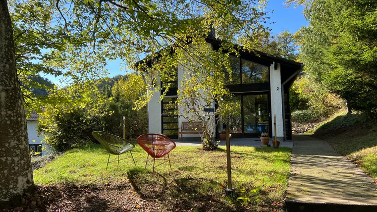 Ein modernes Haus umgeben von Bäumen und grünem Gras. Vor dem Haus stehen zwei gemütliche Stühle.