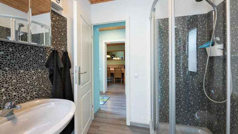 Een moderne badkamer met een douchecabine en een wastafel. Op de achtergrond is een aangrenzende ruimte met tafels en stoelen zichtbaar.