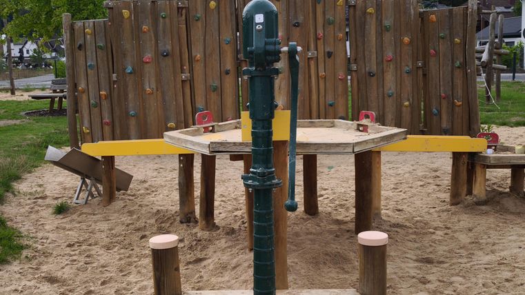 Een speeltuin met een klimwand en een waterspeeltoestel ligt in een zanderige omgeving. Op de achtergrond zijn groene bomen en een woonwijk te zien.
