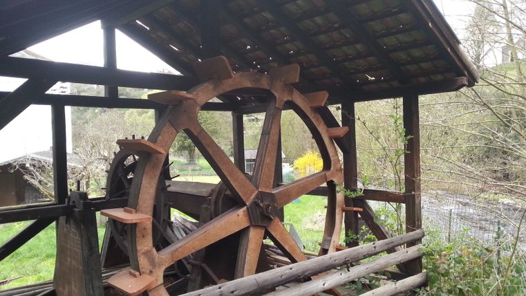 Altes Wasserrad einer Ölmühle mit Holzdach und umgebender Natur.
