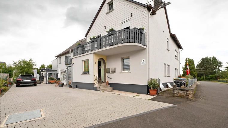 Ein modernes Haus mit einem überdachten Balkon und einer gepflasterten Einfahrt. Die Umgebung ist grün und gut gepflegt.