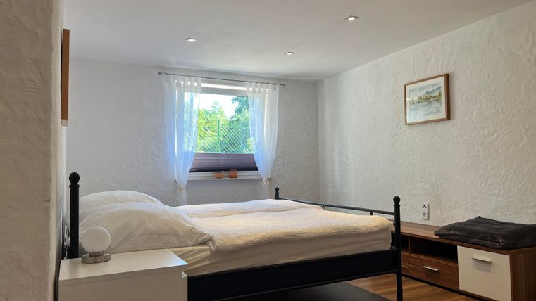 Ein helles Schlafzimmer mit einem Doppelbett und Holzfußboden. Die Fenster sind mit weißen Vorhängen dekoriert und es gibt einen Nachttisch und eine Kommode.