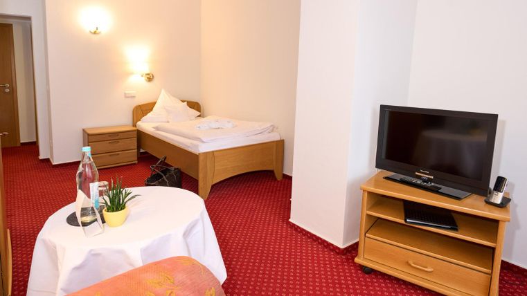 Een eenvoudige hotelkamer met een eenpersoonsbed, een televisie en een klein tafeltje. Het tapijt is rood en de muren zijn licht ontworpen.