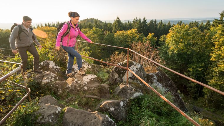 Twee wandelaars op een rotsachtig pad met leuningen, omgeven door bos en uitzicht op heuvels.