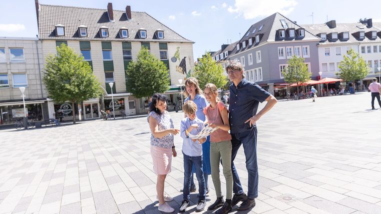 Eine Gruppe von fünf Personen steht auf einem Platz. Im Hintergrund sind einige Gebäude und Bäume zu sehen.