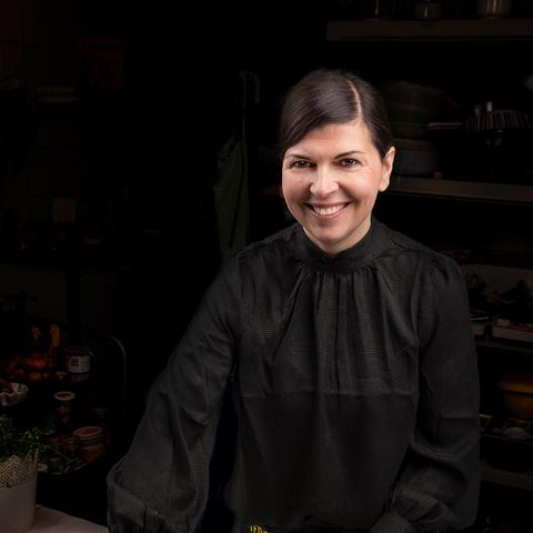 Een glimlachende vrouw in een zwarte blouse staat in een gezellige omgeving. De achtergrond toont verschillende keukengerei en planten.