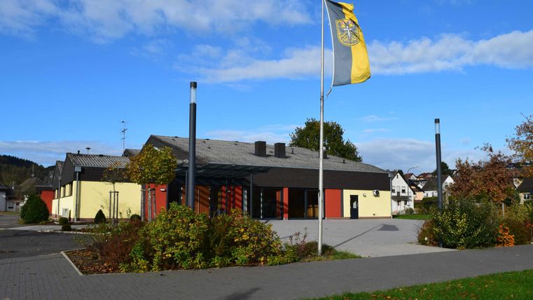 Ein modernes Gebäude mit gelber und schwarzer Fassade. Im Vordergrund stehen Bäume und ein Fahnenmast mit einer gelben Fahne.