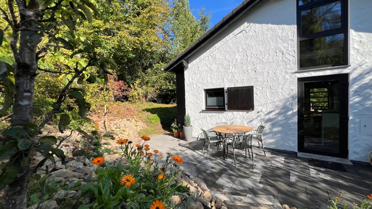 Ein gemütliches Haus mit einer hellen Fassade und großen Fenstern. Vor dem Haus stehen ein Tisch und Stühle sowie bunte Blumen in einem Garten.