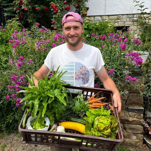 Een lachende man houdt een mand met verse groenten en kruiden vast. Op de achtergrond bloeien kleurrijke bloemen.