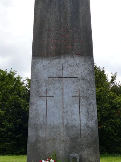 Een gedenksteen met drie kruissymbolen en een inscriptie. Voor de steen staan bloemen en een klein lantaarntje.