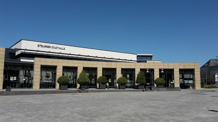 Un bâtiment moderne avec de grandes fenêtres et un ciel bleu clair. Devant le bâtiment se trouvent des arbres disposés régulièrement et des pavés.