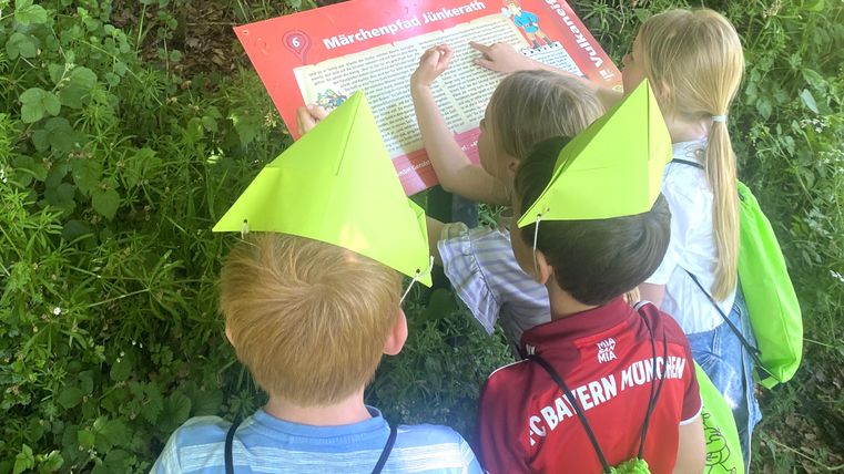 Kinder mit grünen Hüten lesen ein Schild auf dem Märchenpfad Jünkerath.