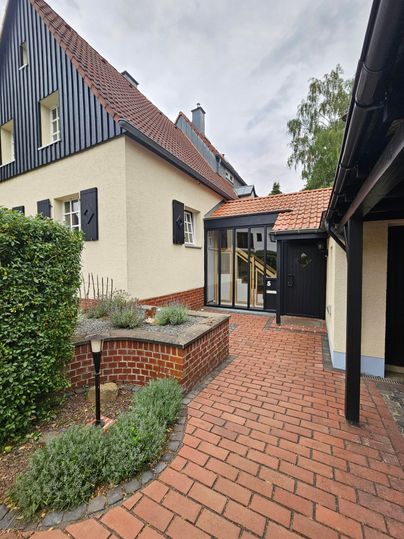 Een uitnodigende buitenruimte van een huis met klinkers en een kleine tuin. De gevel is in twee kleuren vormgegeven, en grote ramen zorgen voor veel licht.
