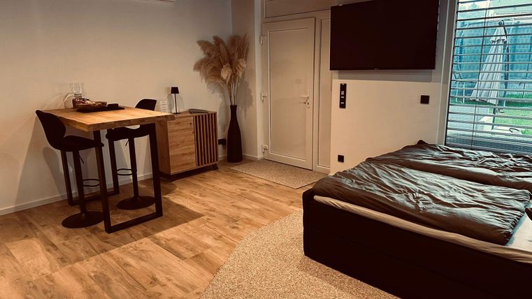 Une chambre moderne avec un lit double, une table en rondins de bois et deux chaises. Il y a de grandes fenêtres qui laissent entrer beaucoup de lumière.