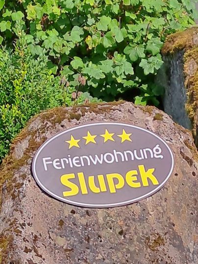 A sign that displays the holiday apartment "Slupek" with four stars. It stands on a large, round stone against a green background.