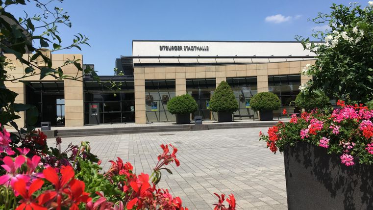 Ein modernes Gebäude mit großen Fenstern und einem klaren blauen Himmel. Bunte Blumen in Töpfen rahmen den Platz ein.