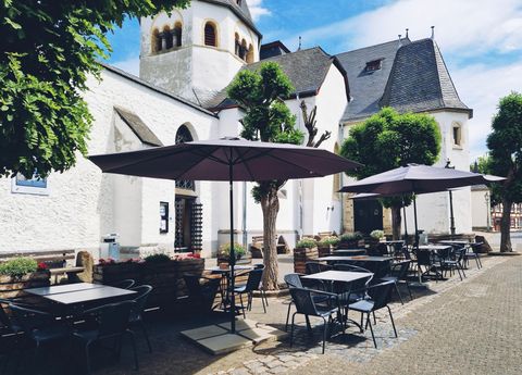 Un café pittoresque avec des tables et des chaises sous des parasols. En arrière-plan, on peut voir une charmante église historique.