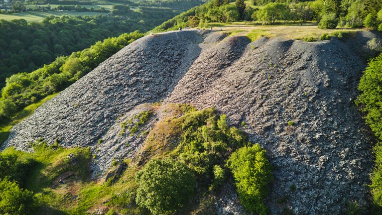 Uitkijkpunt Maria Schacht in de Eifel: Op de voorgrond een grote, grijze leisteenhelling met schaarse begroeiing, omgeven door groene bomen. Daarachter een vlakke weide met bomen, op de achtergrond beboste heuvels en velden onder een heldere hemel.