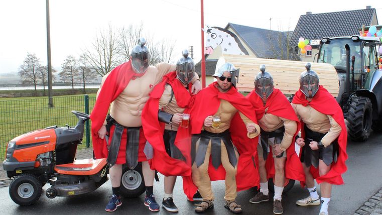 Five men in Roman costumes with helmets and red capes are standing on a street. Next to them is a lawn mower tractor.