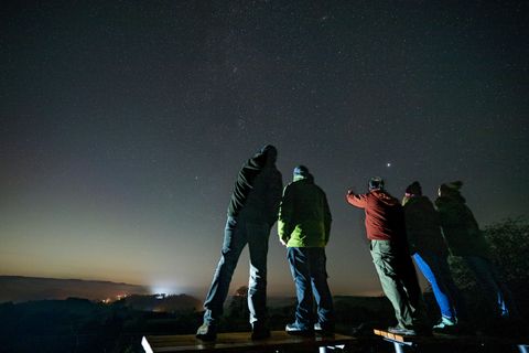 Een groep mensen staat 's nachts onder de sterrenhemel. Ze kijken naar de lucht en wijzen naar de sterren.
