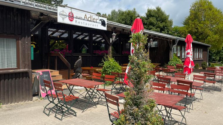 Een restaurant met een buitenruimte en houten meubels. Rode parasols staan op het plein onder een heldere lucht.