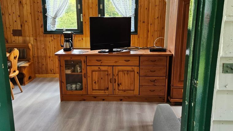 A cozy living space with wood paneling and large windows. A television is on a wooden dresser, and the floors are gray.