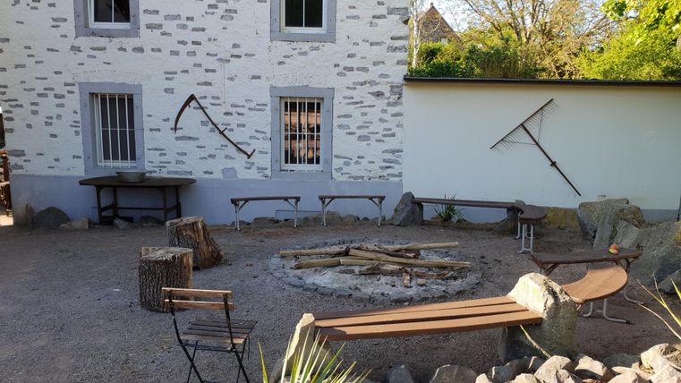 Ein gemütlicher Innenhof mit einem Feuerplatz und umgeben von Bänken. Die Wände sind schlicht und es gibt einige Werkzeuge zur Dekoration.
