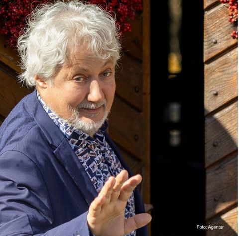 Ein Mann mit grauen Haaren winkt lächelnd in die Kamera. Er trägt einen blauen Anzug und ein gemustertes Hemd.