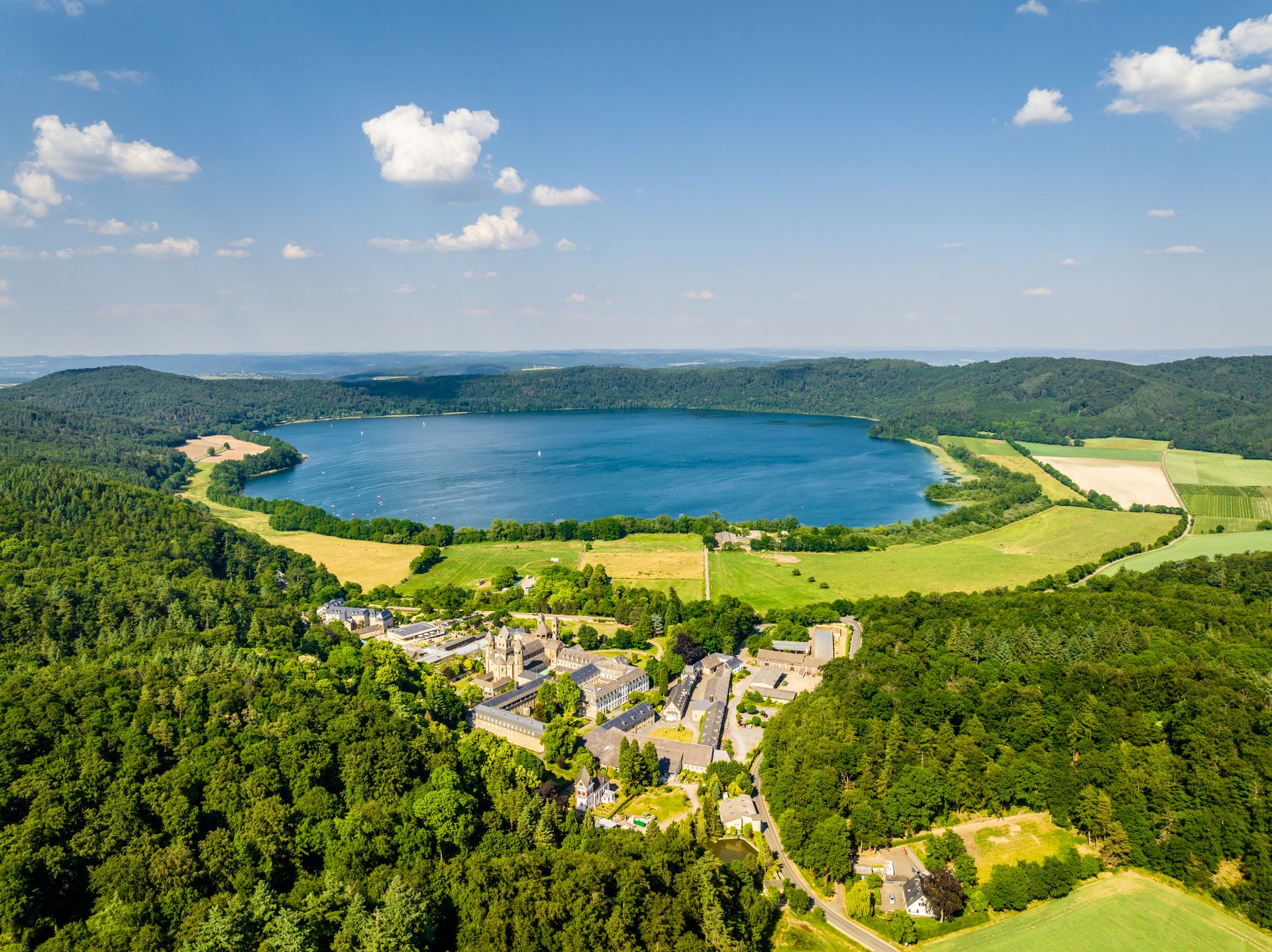 Panorama-Luftaufnahme des Laacher Sees, umgeben von dichten Wäldern und Feldern; im Vordergrund liegt die Abtei Maria Laach mit ihren Gebäuden eingebettet in die grüne Landschaft unter blauem Himmel mit vereinzelten Wolken.