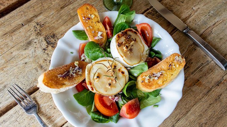 Ein Teller mit frischem Salat, Tomaten und knusprigem Brot. Oben auf dem Salat liegt geschmolzener Käse, der mit Zwiebeln garniert ist.