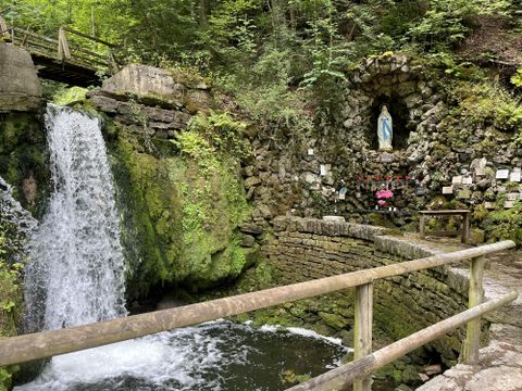 Een schilderachtige bron met waterval, omgeven door groene planten. Aan de rotswand bevindt zich een standbeeld van Maria, omgeven door bloemen.