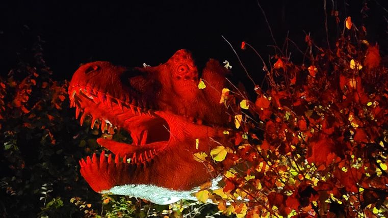 Une tête de dinosaure en plastique brille rouge dans l'obscurité. Autour, on peut voir des feuilles colorées et des accents lumineux.